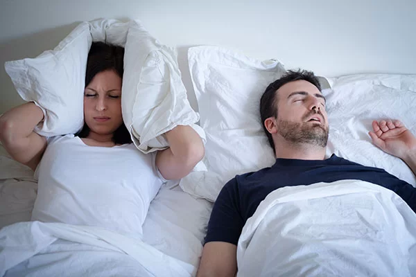 woman covering her ears with her pillow from her bed partner's loud snoring